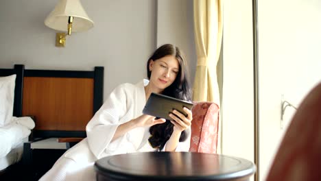 Joven-empresaria-en-bata-de-baño-con-tableta-de-ordenador-sentado-en-la-silla-en-la-habitación-del-hotel