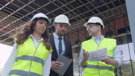 Team-of-Two-Engineers-and-Businessman-in-Hard-Hat-Walking,-Talking,-and-Using-Tablet-Computer.-Glass-Building-or-Skyscraper-under-Construction-on-Background.