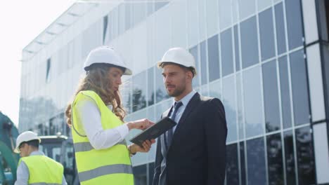 Weibliche-Ingenieur-und-Geschäftsmann-in-harte-Hüte-mit-Unterhaltung-und-Tablet-auf-Baustelle.-Glasbau-auf-Hintergrund.