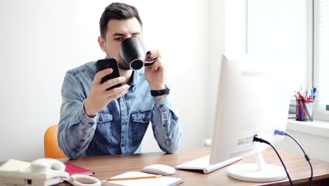 Junge-Büroangestellte-mit-seinem-Telefon-im-Büro-sitzen-am-Tisch-mit-Computer,-Telefon-und-trinken-Kaffee-oder-Tee-aus-seinem-Becher.-Schuss-in-4k