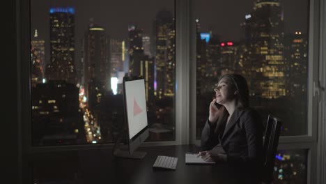 Good-Looking-Woman-Working-with-a-Computer,-Ballpoint-Pen,-Pad-and-a-Phone-in-Multistory-Office-Building.-Calling-Colleagues,-Big-City-Can-Be-Seen-through-Window.