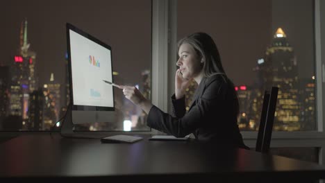 Atractiva-morena-trabajando-en-la-tabla-de-la-oficina-por-la-noche.-Empresaria-de-trabajo-con-ordenador-y-teléfono-inteligente-oficina-con-vista-al-paisaje-urbano.