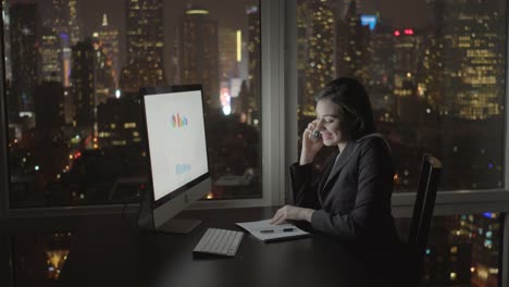 Attraktive-Brünette-am-Bürotisch-nachts-arbeiten.-Geschäftsfrau,-die-Arbeit-mit-Computer-und-Smartphone-mit-Stadtbild-Ansicht-im-Amt.