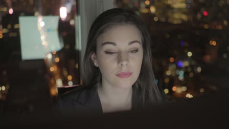 Attractive-Brunette-Working-at-Office-Table-at-Night.-Businesswoman-Working-with-Computer-and-Smartphone-in-Office-with-Cityscape-View.