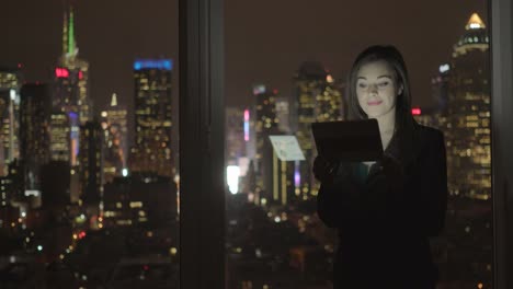 Attractive-Brunette-Working-in-Office-at-Night.-Businesswoman-Working-with-Digital-Tablet-in-Office-with-Cityscape-View.