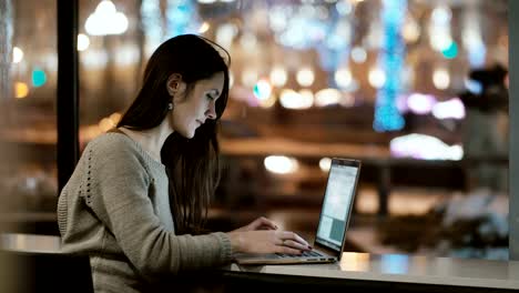 Young-beautiful-woman-sitting-near-the-window-in-the-evening-and-working-on-laptop-computer.-Girl-surfing-the-Internet