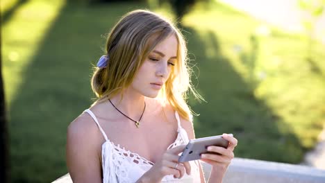 nachdenklich-Blondine-mit-hellen-Augen,-mit-Blick-auf-Handy,-Bildschirm-im-Park,-sonnigen-Sommerwetter-zu-berühren