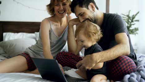 Junge-Familie-mit-niedliche-kleine-Mädchen-spielen-mit-Tochter-während-sie-tippen-auf-Laptop-Computer-sitzen-im-Bett-zu-Hause