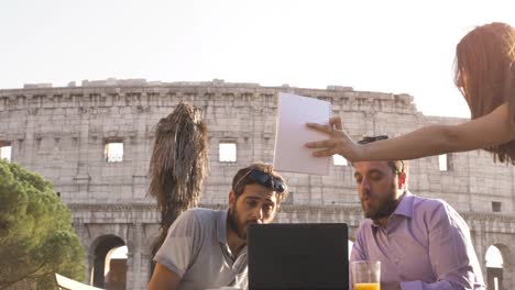 Drei-junge-Menschen,-die-Arbeit-Videocall-mit-Laptop-arbeiten-außerhalb-pitching-zusammen-ein-Projekt-sitzen-am-Stehtisch-Restaurant-vor-Colosseum-in-Rom-bei-Sonnenuntergang