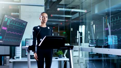 Hermosa-mujer-atleta-con-electrodos-conectados-a-su-cuerpo-camina-en-una-cinta-de-correr-en-un-laboratorio-de-ciencia-de-deportes.-En-el-laboratorio-de-alta-tecnología-de-fondo-con-monitores-mostrando-lecturas-de-electrocardiograma.