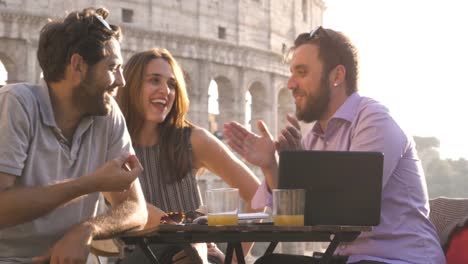 Drei-junge-Freunde,-die-Spaß-mit-Laptop-und-Tablet-sitzen-am-Stehtisch-Restaurant-vor-Colosseum-in-Rom-bei-Sonnenuntergang-zum-Lachen