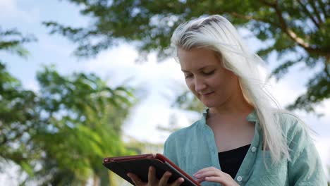 Beautiful-slim-woman-with-long-blonde-hair-in-green-shirt-sits-on-the-ground-and-using-smartphone-over-background-the-park.-Girl-on-the-square-touching-screen-and-smile