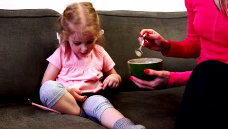 Mom-spoon-feeds-her-daughter-who-looks-at-the-phone