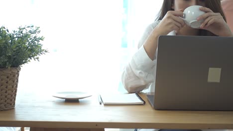 Beautiful-young-smiling-woman-working-on-laptop-while-enjoying-drinking-warm-coffee-sitting-in-a-living-room-at-home.-Enjoying-time-at-home.-Asian-business-woman-working-in-her-home-office.