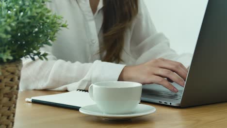 Beautiful-young-smiling-woman-working-on-laptop-while-enjoying-drinking-warm-coffee-sitting-in-a-living-room-at-home.-Enjoying-time-at-home.-Asian-business-woman-working-in-her-home-office.