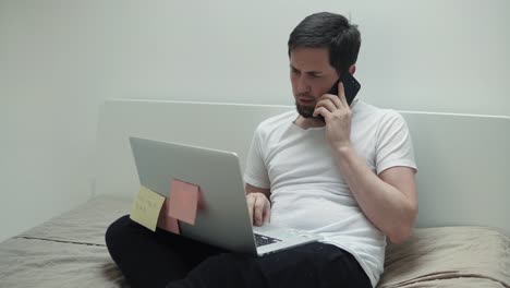 Busy-man-is-talking-by-cell-phone-and-typing-on-laptop,-sitting-in-his-bedroom