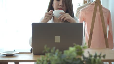 Beautiful-young-smiling-woman-working-on-laptop-while-enjoying-drinking-warm-coffee-sitting-in-a-living-room-at-home.-Enjoying-time-at-home.-Asian-business-woman-working-in-her-home-office.