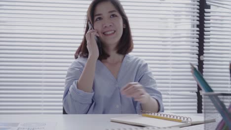 Beautiful-young-smiling-asian-woman-working-on-laptop-while-sitting-in-a-living-room-at-home.-Asian-business-woman-using-phone-for-work-in-her-home-office.-Enjoying-time-at-home.