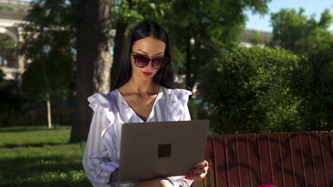 Elegante-morena-en-gafas-de-sol-escribiendo-en-computadora-portátil-sentado-en-un-banco-de-Parque