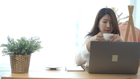 Beautiful-young-smiling-woman-working-on-laptop-while-enjoying-drinking-warm-coffee-sitting-in-a-living-room-at-home.-Enjoying-time-at-home.-Asian-business-woman-working-in-her-home-office.