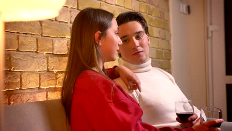 Dolly-close-up-shot-of-young-caucasian-couple-in-profile-sitting-on-sofa-and-drinking-wine-communicating-in-cosy-home-atmosphere.