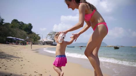 Mamá-e-hija-caminan-a-lo-largo-de-la-costa-a-lo-largo-de-la-arena-en-trajes-de-natación-rosados.-4K