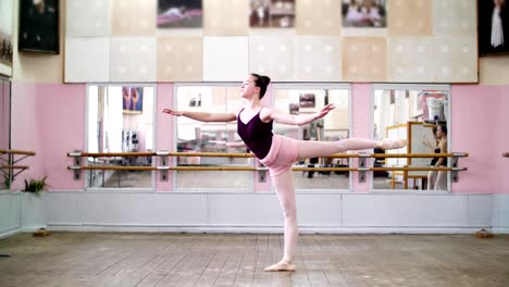 in-dancing-hall,-Young-ballerina-in-black-leotard-performs-1-arabesque,-raises-her-leg-up-behind-elegantly,-standing-near-mirror-in-ballet-class