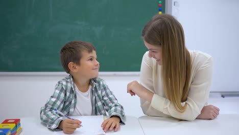 Erfahrenen-Tutor-hilft,-Schüler-am-Tisch-in-der-Nähe-von-Board-im-Klassenzimmer-der-Schule-zu-lernen