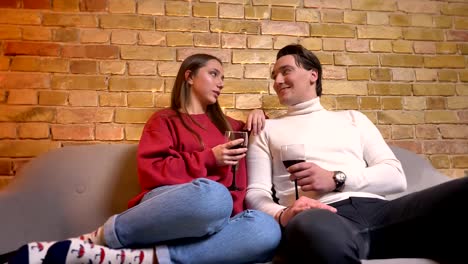 Portrait-of-young-caucasian-couple-sitting-on-sofa-and-drinking-wine-communicating-with-each-other-in-cosy-home-atmosphere.