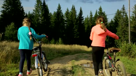 Dos-pequeñas-muchachas-con-bicicletas-paseando-por-la-carretera-rural