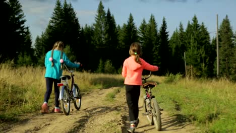 Dos-pequeñas-muchachas-con-bicicletas-paseando-por-la-carretera-rural