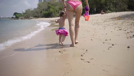 Mom-and-daughter-walk-along-the-seaside-along-the-sand-in-pink-swimsuits.-4K