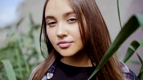 A-young-woman-with-long-hair-stands-amidst-a-tall-grass-and-looks-into-the-camera.