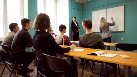 Woman-student-writes-on-a-whiteboard-in-front-of-a-teacher-and-class