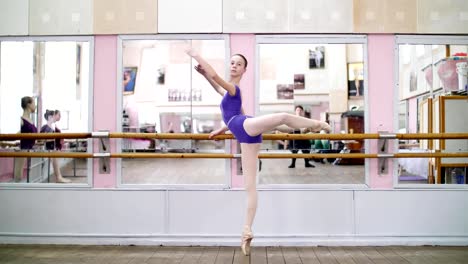 in-dancing-hall,-Young-ballerina-in-purple-leotard-performs-developpe-attitude-on-pointe-shoes,-raises-her-leg-up-behind-elegantly,-standing-near-barre-at-mirror-in-ballet-class