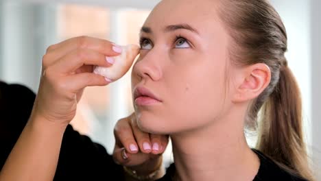 Unrecognizable-visagist-applying-fluid-foundation-on-young-woman's-face-using-sponge.-Professional-makeup-in-salon.-Nude-makeup