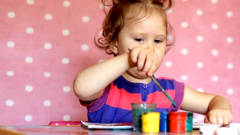 Lustige-kleine-Mädchen-zeichnen-Gouache-an-einem-Tisch-auf-einem-rosa-Hintergrund.-Porträt-von-einem-niedlichen-Baby-mit-einem-Pinsel-und-farbige-Lacke.-Kleine-Künstler.-Kreatives-Konzept.-Kindheit.-Vorschulkind-Bildung