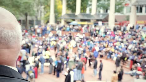 A-older-bold-man-overlooks-a-large-gathering-of-people-in-a-downtown-city