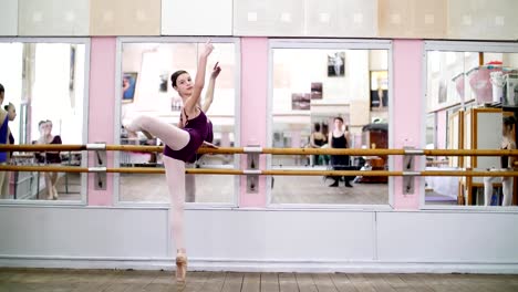 in-dancing-hall,-Young-ballerina-in-purple-leotard-performs-developpe-attitude-on-pointe-shoes,-raises-her-leg-up-behind-elegantly,-standing-near-barre-at-mirror-in-ballet-class