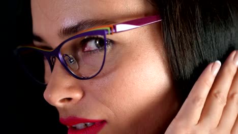 portrait-of-a-sexy-brunette-woman-with-red-lips-who-eroticly-and-playfully-tries-on-stylish-glasses,-spectacles-and-looking-sexually-at-camera-in-studio.-dark-gray-background