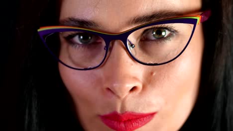 portrait-of-a-sexy-brunette-woman-with-red-lips-who-eroticly-and-playfully-tries-on-stylish-glasses,-spectacles-and-looking-sexually-at-camera-in-studio.-dark-gray-background