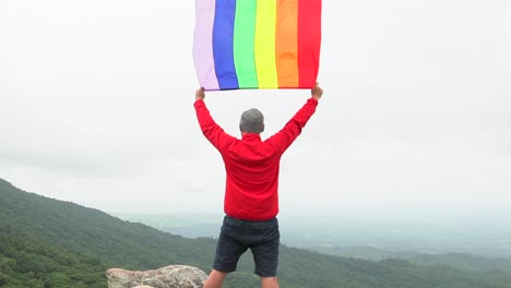 Mann-erhöhen-Regenbogen-Farbe-LGBTI-Fahnenschwingen-in-harten-Wind-auf-Mountain-Top-Sicht