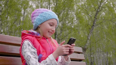 Junges-Mädchen-mit-Telefon-beim-Sitzen-auf-der-Bank-im-Park-begeistert