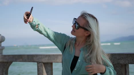 Junge-schöne-schlanke-Frau-mit-langen-blonden-Haaren-mit-Sonnenbrille-und-grünes-Hemd-stehen-in-der-Nähe-von-Palme-und-Selfie-auf-Handy-auf-blauem-Himmel-und-Meer-Hintergrund-zu-machen.-Mädchen-mit-smartphone