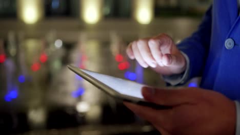 Tracking-shot-of-male-hands-swiping,-zooming-and-clicking-touchscreen-using-tablet-computer-while-man-walking-down-street-at-night