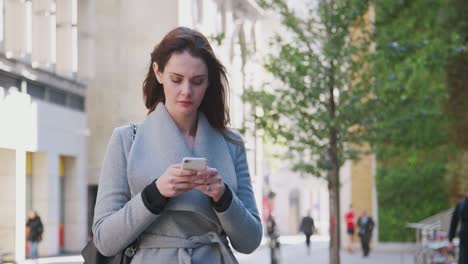 Young-white-woman-walking-in-a-sunny-London-street-stops-and-uses-her-smartphone,-close-up