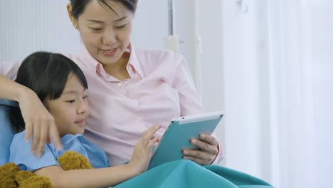 Cute-little-girl-lying-on-bed-with-her-mother-in-hospital,-watching-funny-cartoons,-movies-on-digital-tablet.-illness-and-treatment.