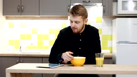 Hombre-joven-con-tableta-comiendo-el-desayuno-sentado-por-la-mesa-en-la-cocina-en-casa.