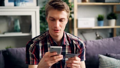 El-hombre-joven-está-haciendo-el-pago-en-línea-con-tarjeta-bancaria-usando-el-teléfono-inteligente-en-casa