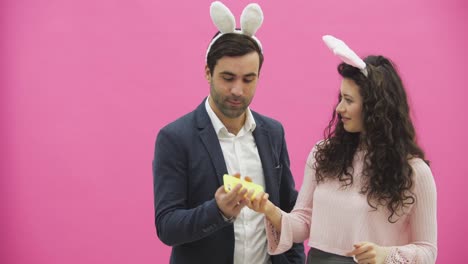 Young-lovers-couple-on-the-pink-background.-With-hackneyed-ears-on-the-head.-During-this,-they-make-a-photo-of-sephi-on-the-phone-showing-various-rabbit-movements-and-rejoice.